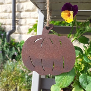 May include: A rusty metal pumpkin cutout hanging from a string. The pumpkin is brown and has a rustic look. It is hanging in front of a potted plant with purple and yellow flowers.