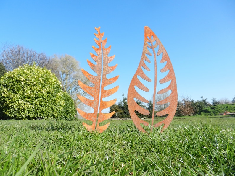 May include: Two rusty metal leaf sculptures in a grassy field. The leaves are cut out in a fern and a simple leaf shape.
