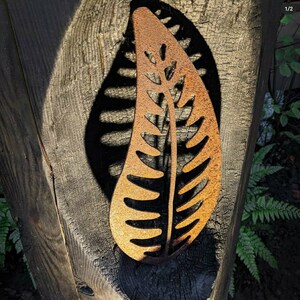 May include: A rusty metal leaf-shaped wall decor with a cutout design. The leaf is attached to a wooden surface.