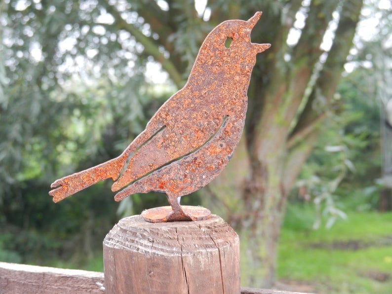May include: A rusty metal bird sculpture with an open beak, perched on a wooden post. The bird has a detailed cut-out design and is set against a blurred green background, suggesting an outdoor setting.