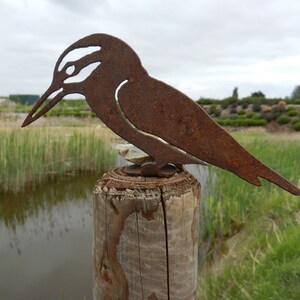May include: A rustic, metal bird sculpture with a weathered, brown finish. The bird is perched on a wooden post, set against a backdrop of a pond and greenery. The sculpture's design features cut-out details.