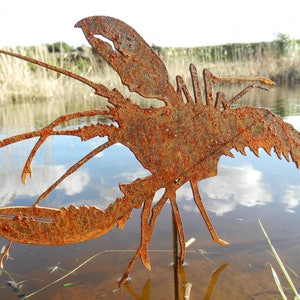May include: A rusty metal lobster sculpture standing in a shallow pond. The sculpture is made of thin metal and is painted in a rusty brown color. The sculpture is standing on a wooden stake.
