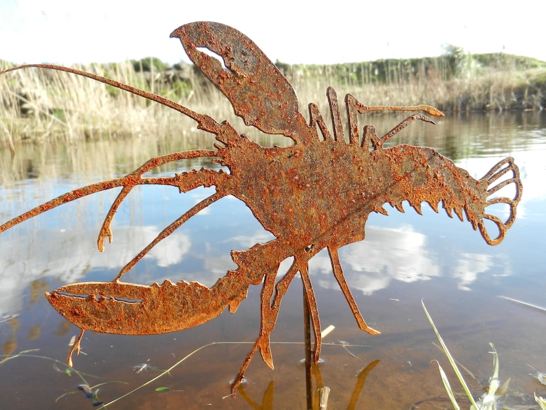 May include: A rusty metal lobster sculpture standing in a shallow pond. The sculpture is made of thin metal and is painted in a rusty brown color. The sculpture is standing on a wooden stake.
