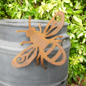 May include: A rusty metal bee cutout with a decorative swirl design on the wings. The bee is attached to a gray planter.