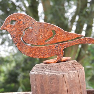 May include: A rustic, metal bird sculpture with a weathered, orange-brown finish. The bird is perched on a wooden post, showcasing a cutout design within its body. The sculpture is an outdoor garden decoration.