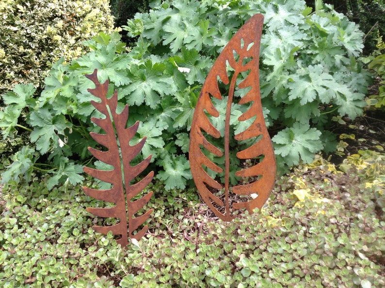 May include: Two rusty metal fern leaf sculptures in a garden setting. The leaves are cut out and have a rustic, weathered look.