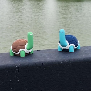 Könnte beinhalten: Zwei kleine, dekorative Schildkrötenfiguren. Eine hat einen braunen Panzer und einen grünen Kopf und Beine, die andere einen blauen Panzer und Kopf. Sie sitzen auf einem schwarzen Geländer mit einem See und Bäumen im Hintergrund.