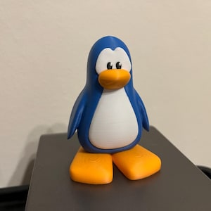 May include: A 3D-printed penguin figurine. The penguin has a blue body, white belly, orange feet and beak, and black eyes. The figurine is standing on a dark gray surface.