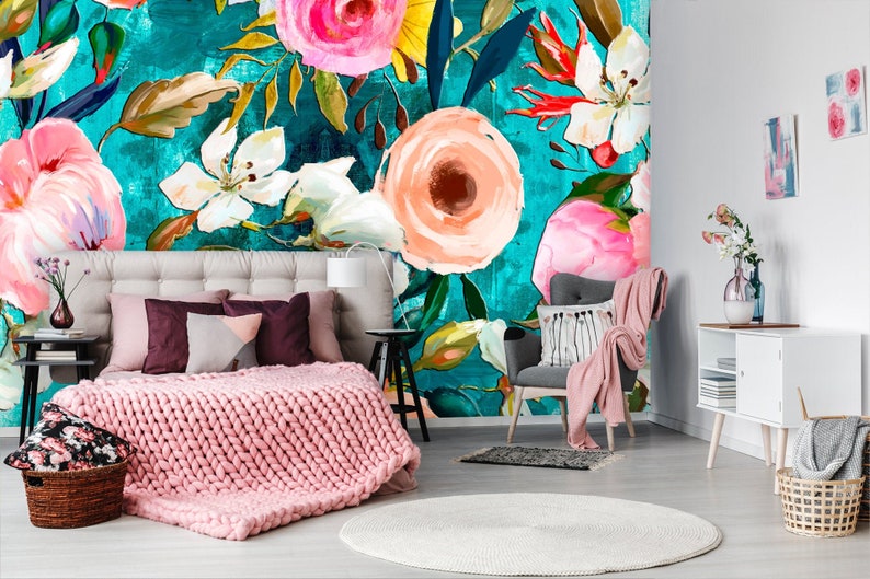 Puede incluir: Un interior de dormitorio con un papel tapiz floral turquesa con flores rosas, blancas y amarillas. La habitaci&oacute;n tiene una cama con una manta de punto rosa, un sill&oacute;n gris y una mesita de noche blanca con un jarr&oacute;n de flores.