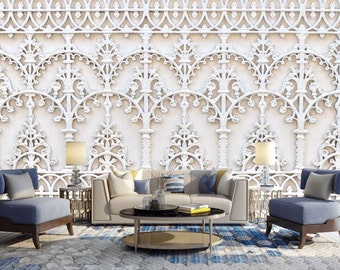 May include: A living room scene featuring a beige sofa, two blue armchairs, and a round coffee table. The room has a patterned rug and is illuminated by lamps. The wall is decorated with a white, ornate architectural design.