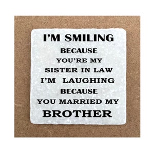 May include: White square coaster with black text that reads "I'm smiling because you're my sister in law. I'm laughing because you married my brother."