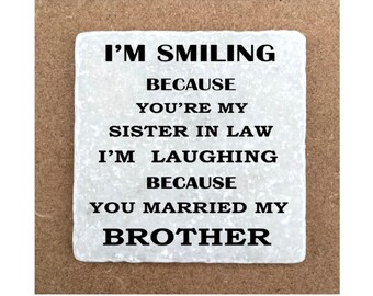 May include: White square coaster with black text that reads "I'm smiling because you're my sister in law. I'm laughing because you married my brother."