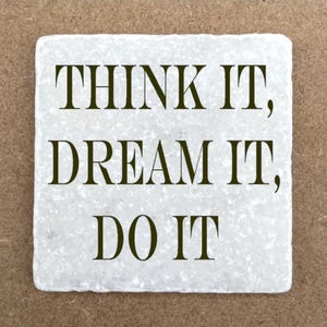 May include: A square, off-white coaster with the words "THINK IT, DREAM IT, DO IT" in dark brown lettering. The coaster has a textured surface and is set against a brown background.
