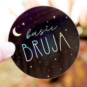 May include: A black and white round sticker with a crescent moon and stars. The text "basic bruja" is written in a rainbow holographic font.
