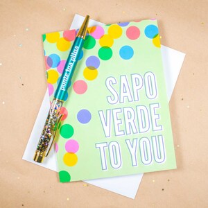 May include: A green card with colorful polka dots and the text "SAPO VERDE TO YOU" in white with a purple outline. A teal pen with gold glitter and the text "ponte las pilas" in gold is on top of the card.