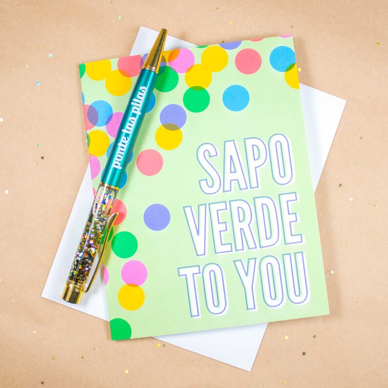 May include: A green card with colorful polka dots and the text "SAPO VERDE TO YOU" in white with a purple outline. A teal pen with gold glitter and the text "ponte las pilas" in gold is on top of the card.
