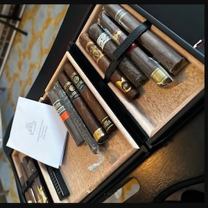 May include: A black, wooden cigar box with a variety of cigars inside. The cigars are various shades of brown and are secured with a black leather strap. A white card with text is visible.