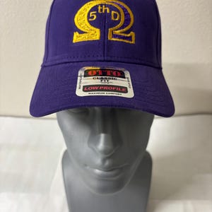Op de afbeelding: Een paarse baseballcap met gouden borduursel. De tekst "5th D" en een Grieks symbool zijn zichtbaar. De cap heeft een gebogen klep en een label met "OTTO CLASSIC FIT LOW PROFILE MAXIMUM COMFORT".
