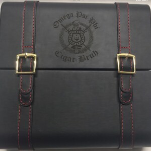 May include: Black leather cigar box with red stitching and gold-colored buckles. The box features the Omega Psi Phi fraternity crest and the words "Cigar Bruh" engraved on the lid. A stylish accessory for cigar enthusiasts.