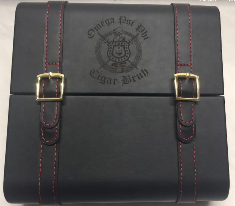 May include: Black leather cigar box with red stitching and gold-colored buckles. The box features the Omega Psi Phi fraternity crest and the words "Cigar Bruh" engraved on the lid. A stylish accessory for cigar enthusiasts.