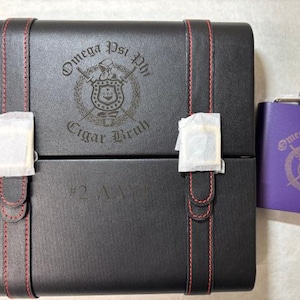May include: A black box with red stitching and the words "Omega Psi Phi Cigar Bruh" and a purple flask with the same logo. The box has a paper wrapping on the front.