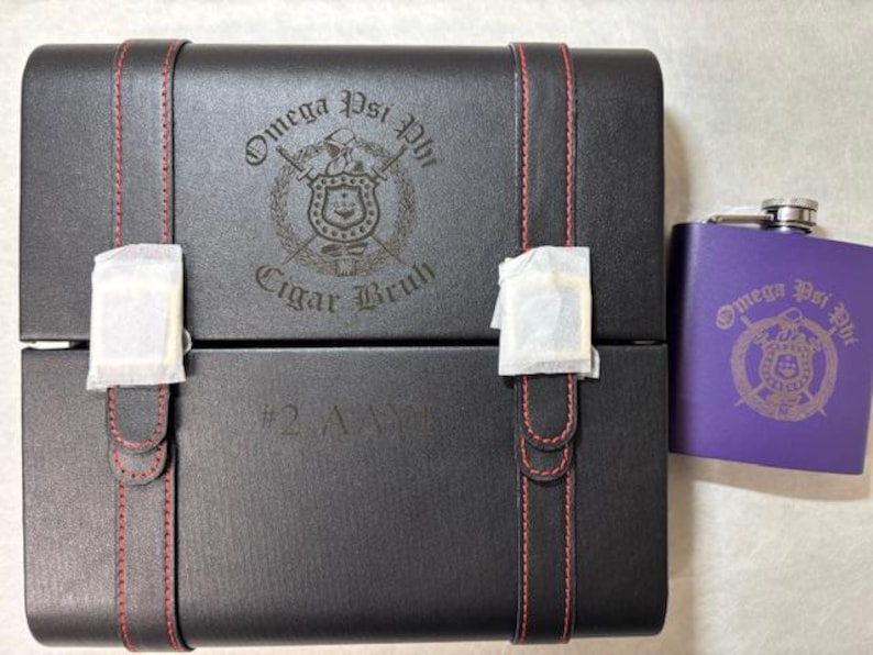 May include: A black box with red stitching and the words "Omega Psi Phi Cigar Bruh" and a purple flask with the same logo. The box has a paper wrapping on the front.