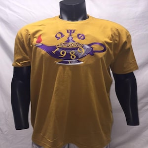 May include: Mustard yellow t-shirt with a purple and gold embroidered genie lamp design. The lamp features the year "1989" and Greek letters. The flame is red. The shirt is displayed on a black mannequin.
