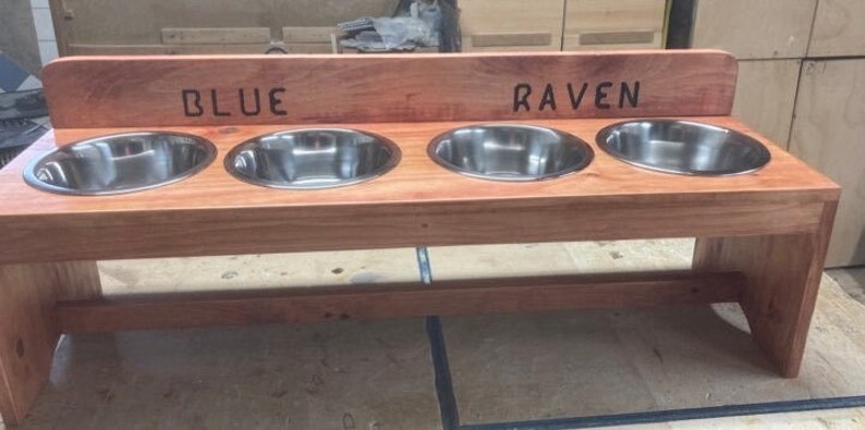 Puede incluir: Un comedero de madera para mascotas con cuatro cuencos de acero inoxidable. El comedero es de madera de color marr&oacute;n rojizo y tiene las palabras "BLUE" y "RAVEN" talladas en la parte superior. Los cuencos est&aacute;n espaciados uniformemente.