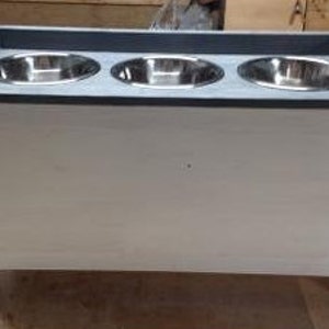 Pu&ograve; includere: Un alimentatore per animali domestici in legno bianco con un piano grigio e tre ciotole in acciaio inossidabile. L'alimentatore &egrave; rettangolare con un frontale in stile armadio ed &egrave; rialzato su gambe corte. Le ciotole sono distanziate uniformemente e incassate nella parte superiore.