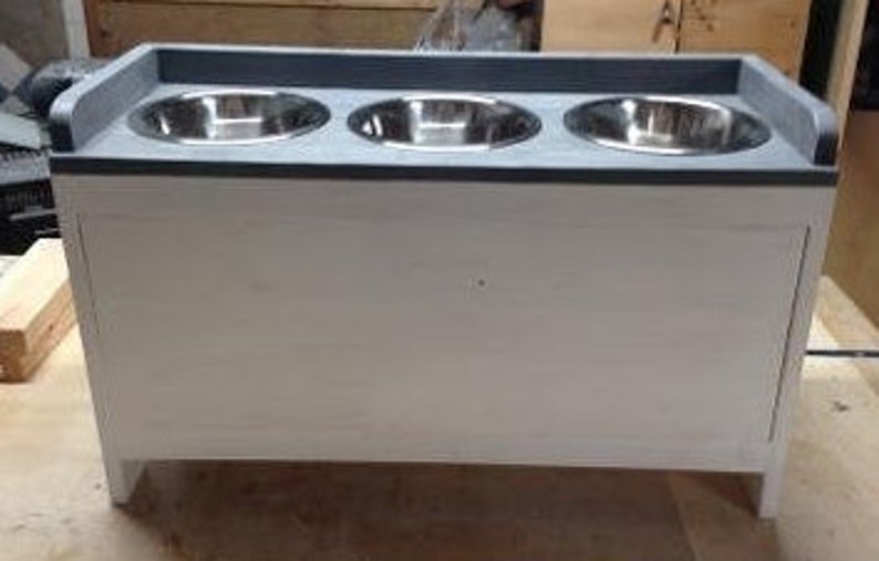 Pu&ograve; includere: Un alimentatore per animali domestici in legno bianco con un piano grigio e tre ciotole in acciaio inossidabile. L'alimentatore &egrave; rettangolare con un frontale in stile armadio ed &egrave; rialzato su gambe corte. Le ciotole sono distanziate uniformemente e incassate nella parte superiore.