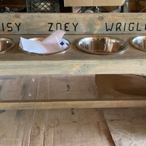 Puede incluir: Un soporte de madera para cuencos para perros con cuatro cuencos de acero inoxidable. Los nombres "DAISY", "ZOEY" y "WRIGLEY" est&aacute;n tallados en la parte superior. El soporte es de madera gris claro.