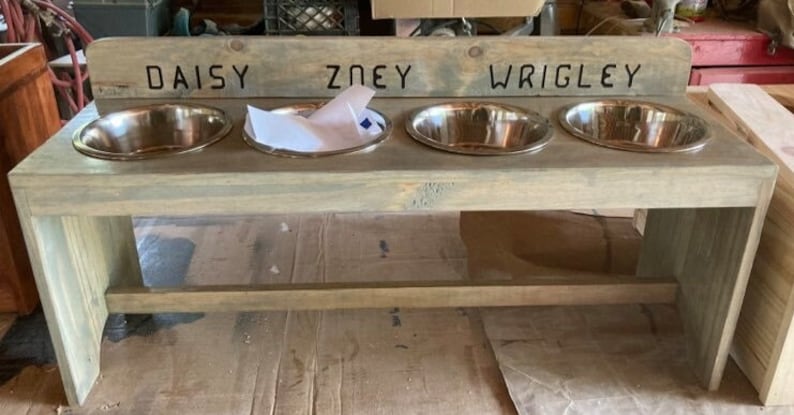 Puede incluir: Un soporte de madera para cuencos para perros con cuatro cuencos de acero inoxidable. Los nombres "DAISY", "ZOEY" y "WRIGLEY" est&aacute;n tallados en la parte superior. El soporte es de madera gris claro.