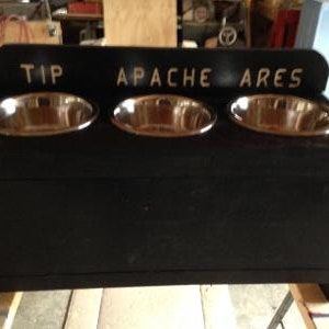 Pu&ograve; includere: Un alimentatore per animali domestici in legno nero con tre ciotole in acciaio inossidabile. La parte superiore dell'alimentatore riporta le parole "TIP APACHE ARES" in lettere bianche. Le ciotole sono di forma ovale e inserite nella struttura in legno.