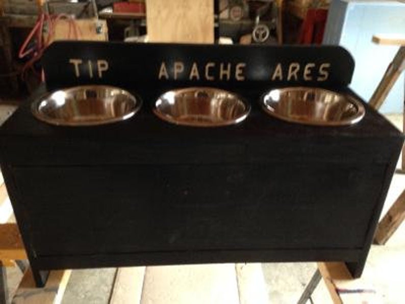 Pu&ograve; includere: Un alimentatore per animali domestici in legno nero con tre ciotole in acciaio inossidabile. La parte superiore dell'alimentatore riporta le parole "TIP APACHE ARES" in lettere bianche. Le ciotole sono di forma ovale e inserite nella struttura in legno.