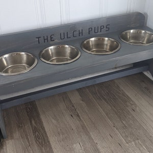 Puede incluir: Un soporte de madera gris para cuencos de perro con cuatro cuencos de acero inoxidable. El texto "THE ULCH PUPS" est&aacute; escrito en la parte superior. El soporte est&aacute; colocado sobre un suelo con aspecto de madera y contra una pared blanca.