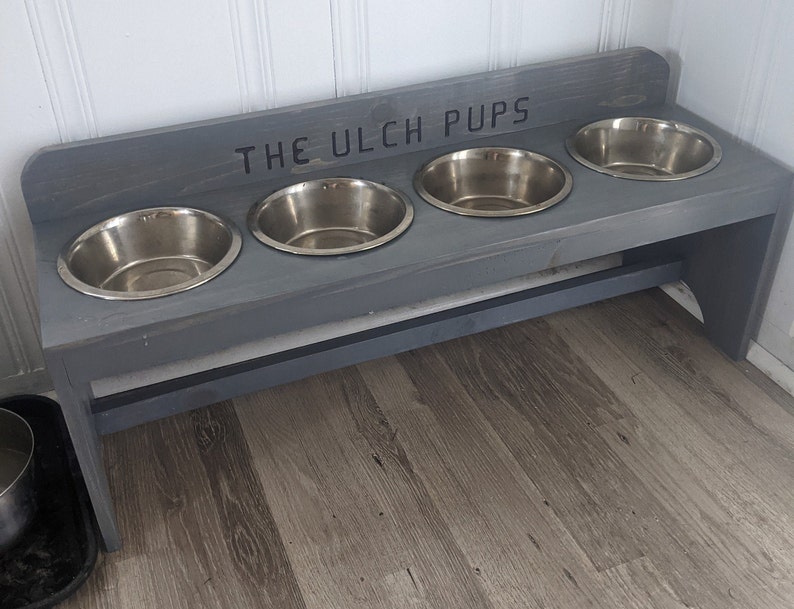 Puede incluir: Un soporte de madera gris para cuencos de perro con cuatro cuencos de acero inoxidable. El texto "THE ULCH PUPS" est&aacute; escrito en la parte superior. El soporte est&aacute; colocado sobre un suelo con aspecto de madera y contra una pared blanca.