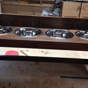 Puede incluir: Un comedero para perros de madera con cuatro cuencos de acero inoxidable. El comedero de madera te&ntilde;ida oscura tiene los nombres "LAILA DIESEL SASHA" grabados en la parte superior. El comedero est&aacute; elevado sobre patas y tiene una barra de soporte negra.