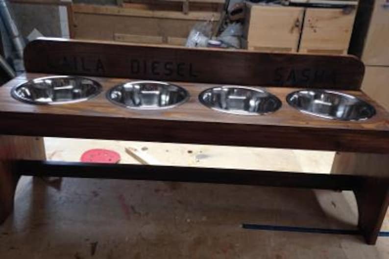 Puede incluir: Un comedero para perros de madera con cuatro cuencos de acero inoxidable. El comedero de madera te&ntilde;ida oscura tiene los nombres "LAILA DIESEL SASHA" grabados en la parte superior. El comedero est&aacute; elevado sobre patas y tiene una barra de soporte negra.