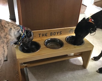 May include: A wooden dog bowl stand with three stainless steel bowls. The stand has the text "THE BOYS" and a paw print. Two dogs are eating from the bowls. The dogs are wearing colorful collars.