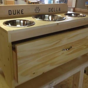 Pu&ograve; includere: Una mangiatoia in legno per animali domestici con tre ciotole in acciaio inossidabile. La sezione superiore ha i nomi "DUKE" e "BELLA" con un'impronta di zampa. Un cassetto &egrave; sotto le ciotole. La mangiatoia &egrave; in legno chiaro.