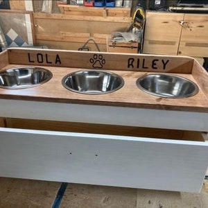 Pu&ograve; includere: Una mangiatoia in legno per animali domestici con tre ciotole in acciaio inossidabile. La parte superiore &egrave; in legno tinto con i nomi "LOLA" e "RILEY" e un'impronta di zampa. La base &egrave; bianca con un cassetto.