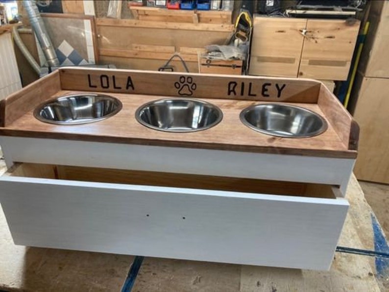 Pu&ograve; includere: Una mangiatoia in legno per animali domestici con tre ciotole in acciaio inossidabile. La parte superiore &egrave; in legno tinto con i nomi "LOLA" e "RILEY" e un'impronta di zampa. La base &egrave; bianca con un cassetto.