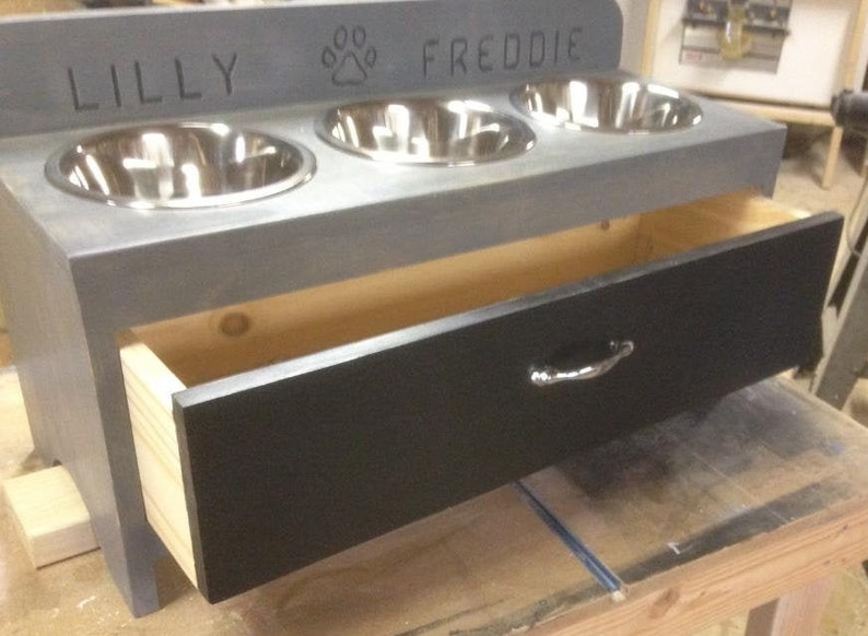 Pu&ograve; includere: Un alimentatore per animali domestici in legno grigio con tre ciotole in acciaio inossidabile e un cassetto estraibile. I nomi "LILLY" e "FREDDIE" sono incisi sopra le ciotole, con un simbolo di impronta di zampa in mezzo. Il cassetto ha una maniglia argentata.