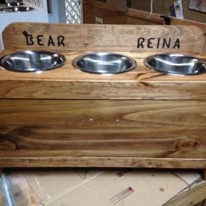 Pu&ograve; includere: Un alimentatore per animali domestici in legno con tre ciotole in acciaio inossidabile. L'alimentatore &egrave; tinto di un marrone caldo e presenta i nomi "BEAR" e "REINA" incisi sulla parte superiore. Le ciotole sono incassate.
