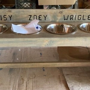 Puede incluir: Un comedero de madera gris claro para mascotas con cuatro cuencos de acero inoxidable. Los nombres "DAISY", "ZOEY" y "WRIGLEY" est&aacute;n tallados en la parte superior. Los cuencos ovalados est&aacute;n encajados en el marco de madera.