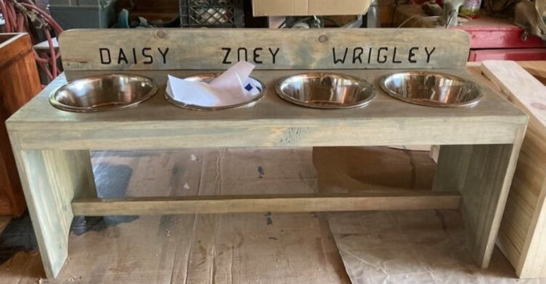 Puede incluir: Un comedero de madera gris claro para mascotas con cuatro cuencos de acero inoxidable. Los nombres "DAISY", "ZOEY" y "WRIGLEY" est&aacute;n tallados en la parte superior. Los cuencos ovalados est&aacute;n encajados en el marco de madera.