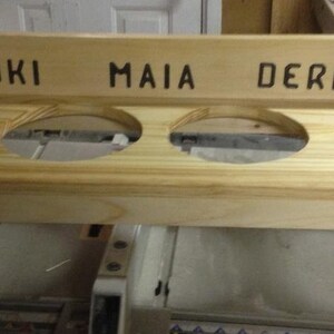 Puede incluir: Un comedero de madera para mascotas con cuatro aberturas circulares para cuencos. La secci&oacute;n superior est&aacute; grabada con los nombres "LOKI", "MAIA" y "DERPY", junto con dise&ntilde;os de huellas de patas. El comedero es de madera clara.