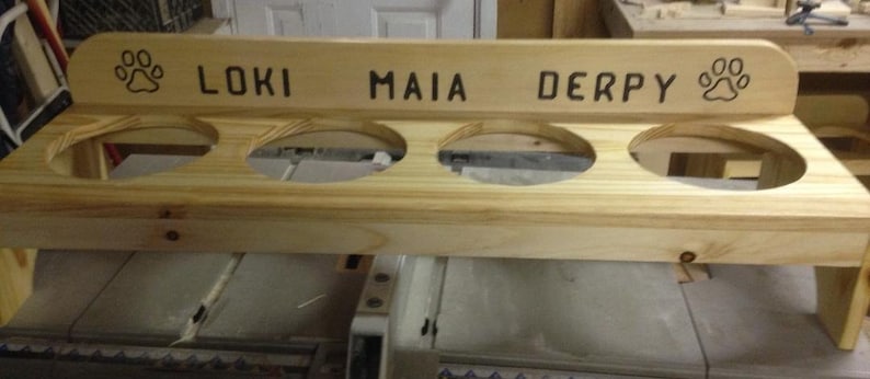 Puede incluir: Un comedero de madera para mascotas con cuatro aberturas circulares para cuencos. La secci&oacute;n superior est&aacute; grabada con los nombres "LOKI", "MAIA" y "DERPY", junto con dise&ntilde;os de huellas de patas. El comedero es de madera clara.