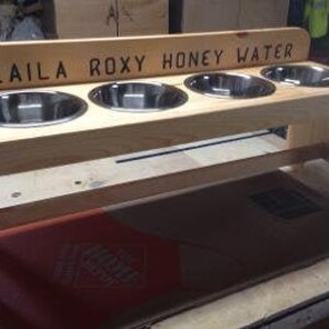 Puede incluir: Un comedero de madera para mascotas con cuatro cuencos de acero inoxidable. El comedero tiene un acabado marr&oacute;n claro y el texto "LAILA ROXY HONEY WATER" est&aacute; grabado en la parte superior. Dise&ntilde;ado para comida y agua.