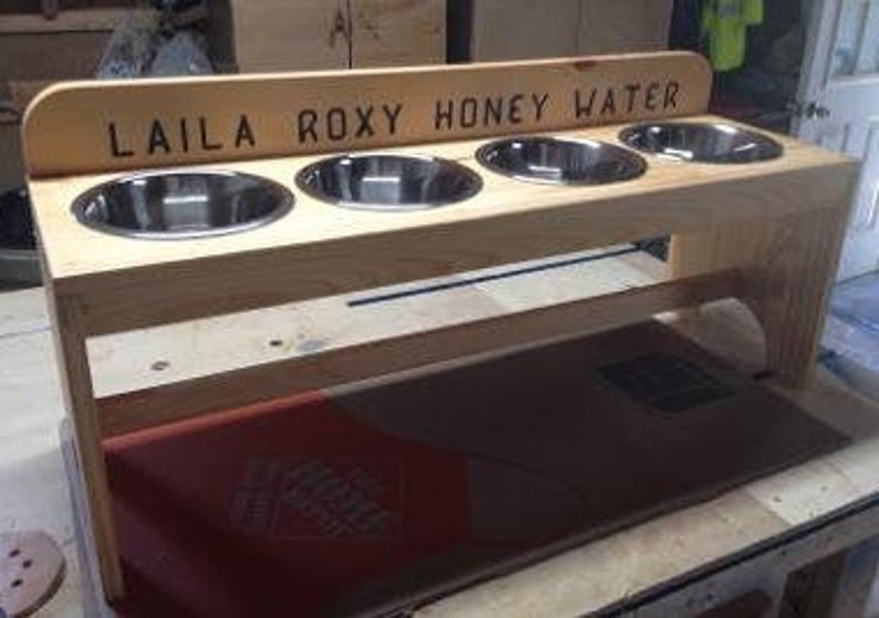 Puede incluir: Un comedero de madera para mascotas con cuatro cuencos de acero inoxidable. El comedero tiene un acabado marr&oacute;n claro y el texto "LAILA ROXY HONEY WATER" est&aacute; grabado en la parte superior. Dise&ntilde;ado para comida y agua.